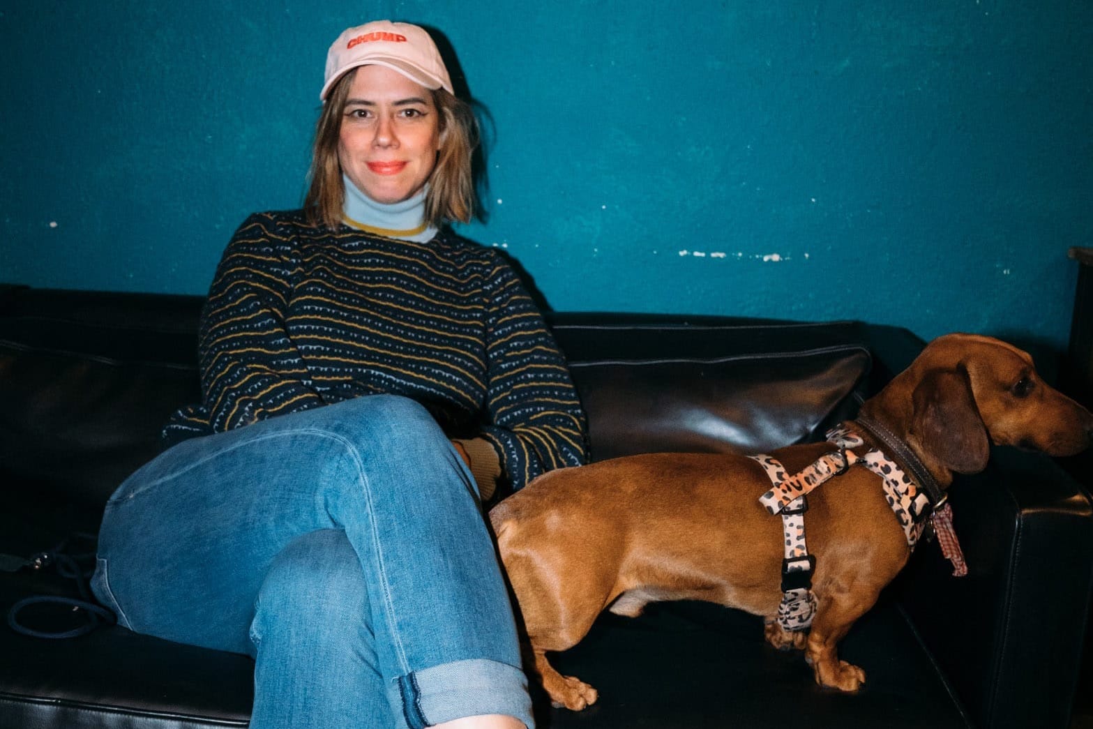 A woman wearing a pink cap, jumper and jeans sits on a sofa next to a sausage dog