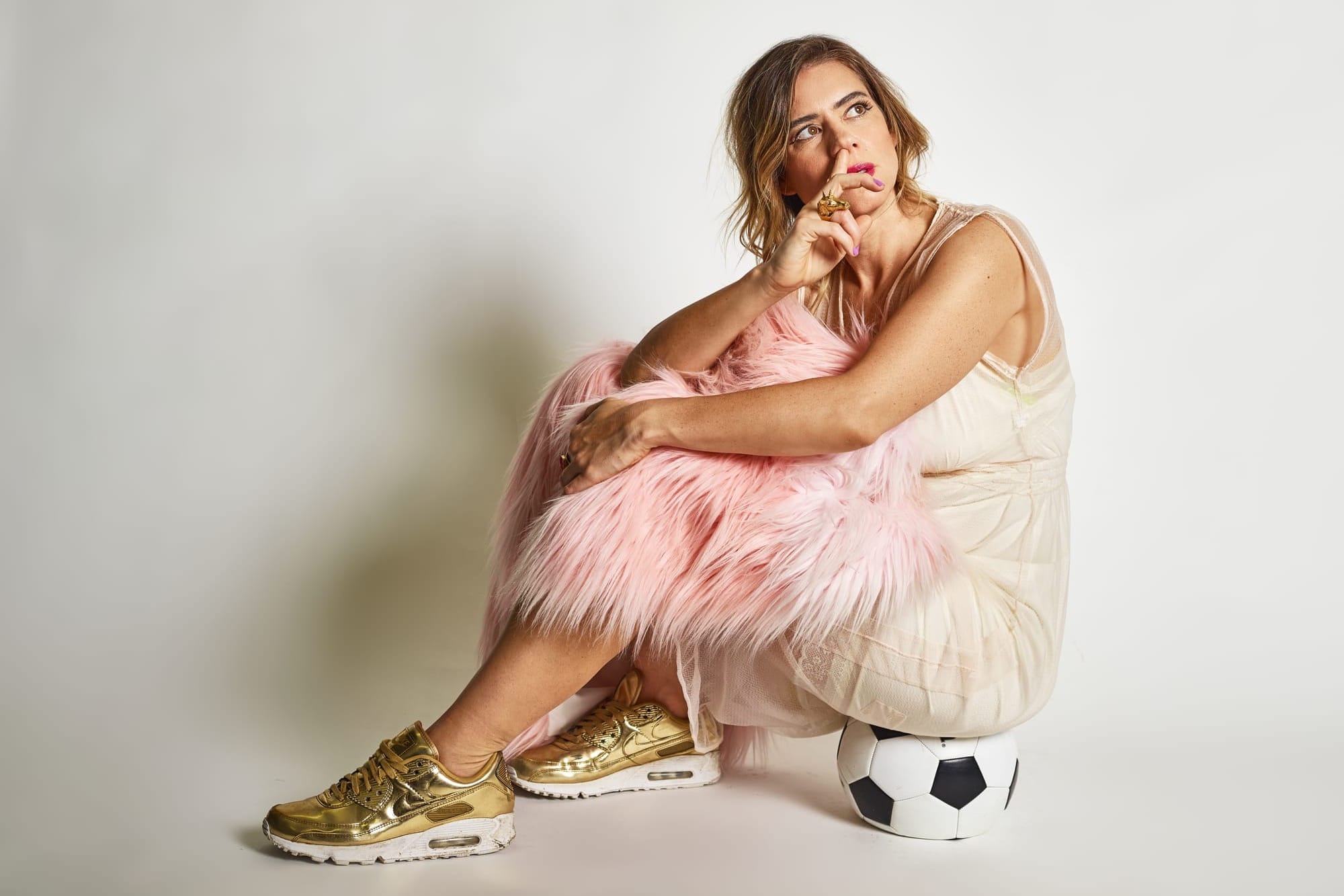 A woman with long brown hair wearing a cream dress and gold trainers sits on a football while picking her nose