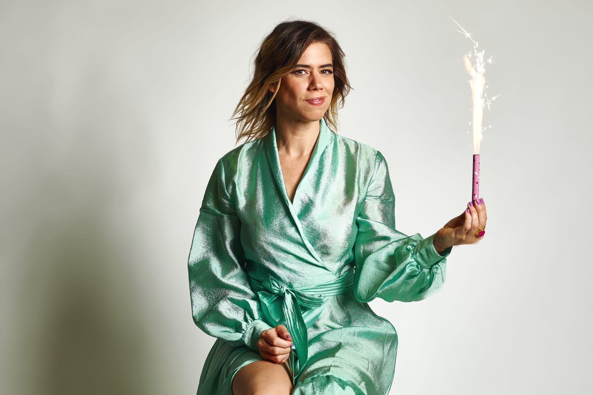A woman with long brown hair wearing a green ball gown holds a firework