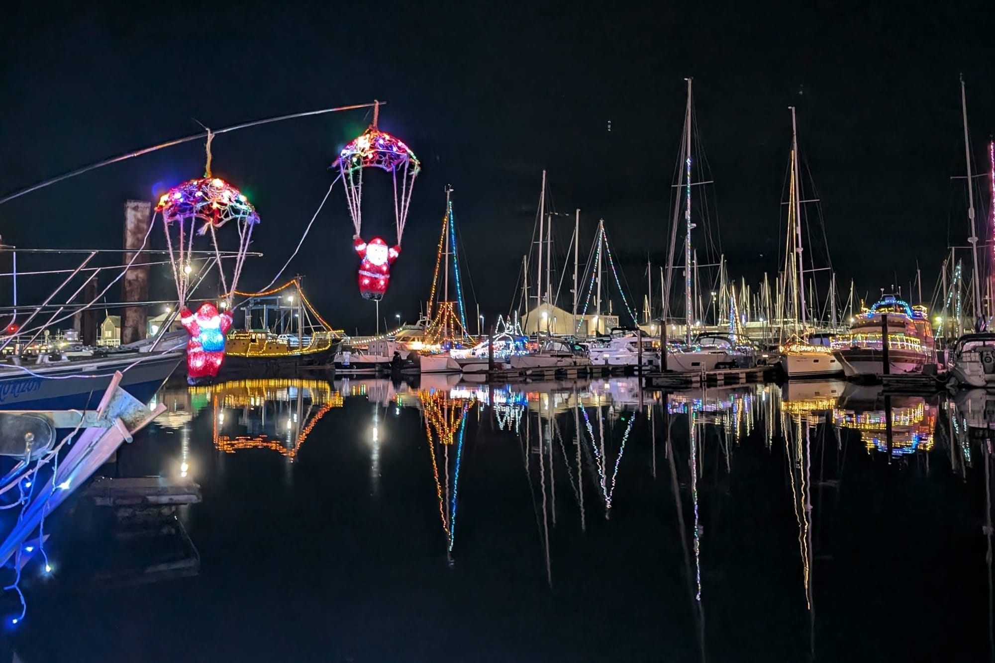 Illuminated boats in a harbour. Two Santas appear to parachute in