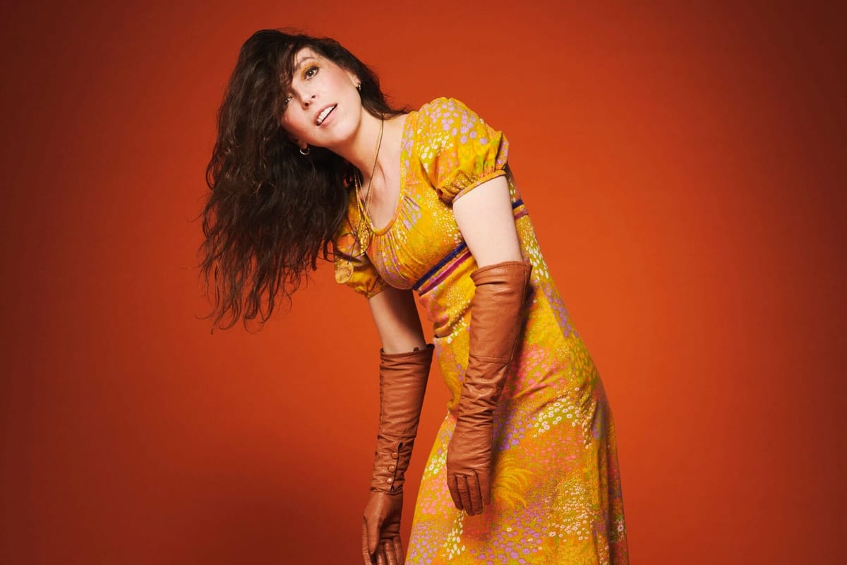 A brunette woman in an orange dress and gloves leans to the side