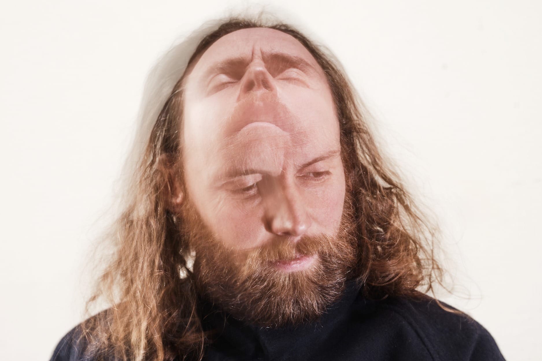 A blurred photo of a man with long brown hair and a beard. His face appears in two positions - looking up with his eyes closed and down to the side