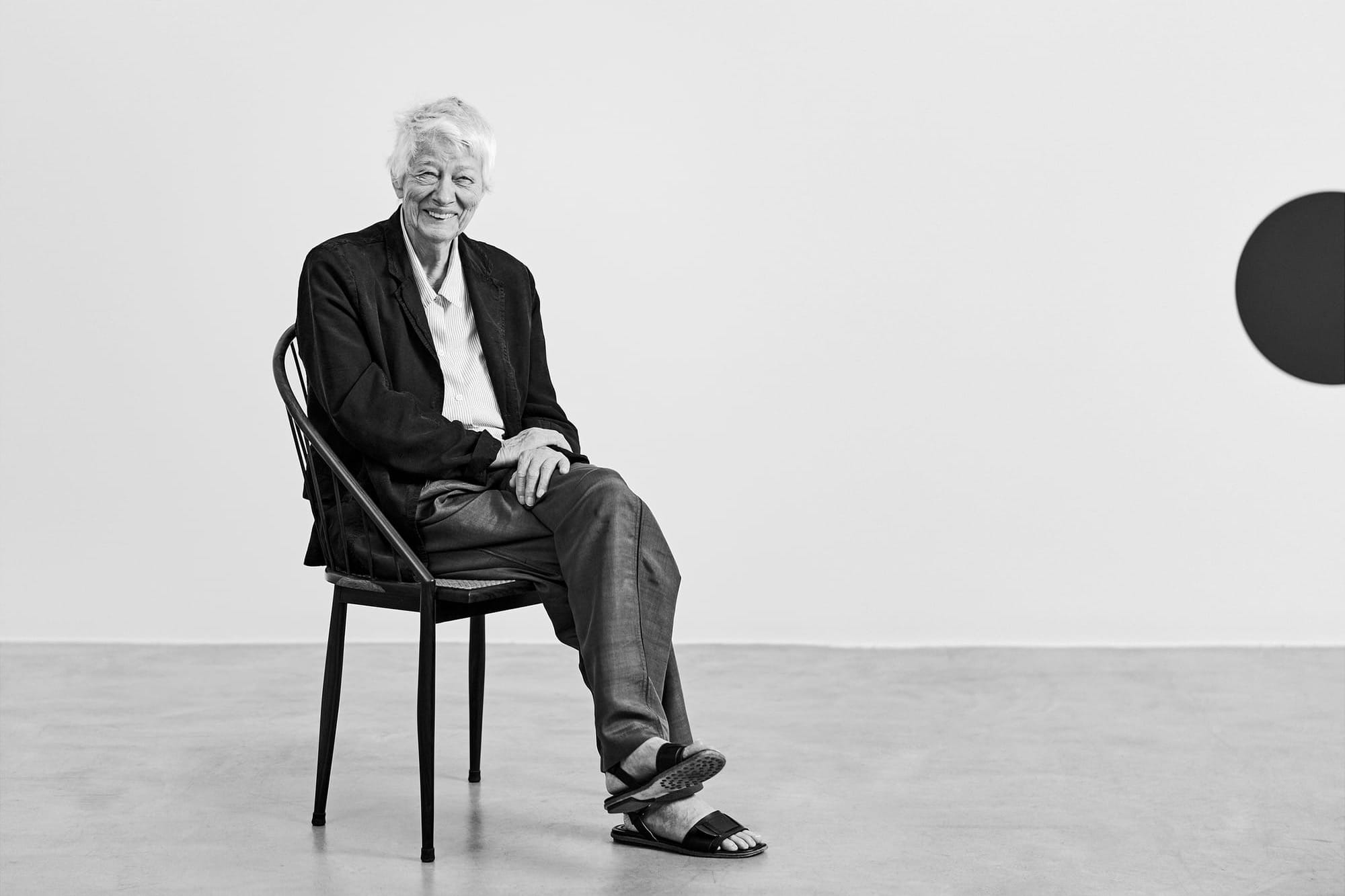 A woman in her 90s with cropped white hair wearing a casual suit sits on a black wooden chair in an art gallery