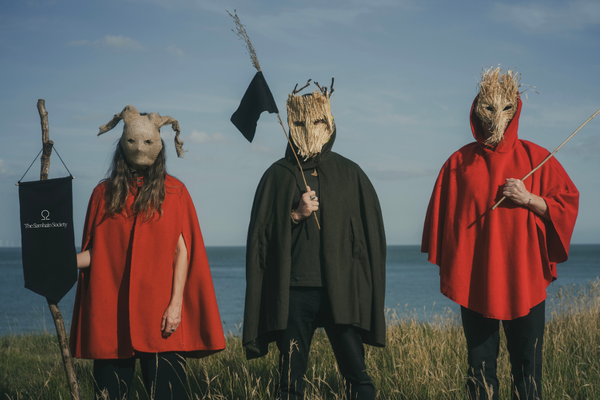 Three people stand on a cliff top in robes and masks carrying flags