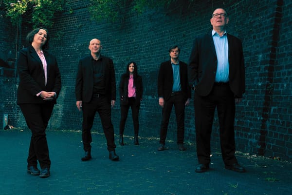 Five people in black suits stand in front of a wall